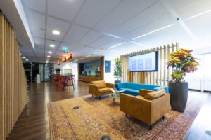Moderne kantoorlobby aan de Johan Huizingalaan met zitgedeelte, grote planten, flatscreen en een koffiebar met hoge tafels en rode barkrukken.